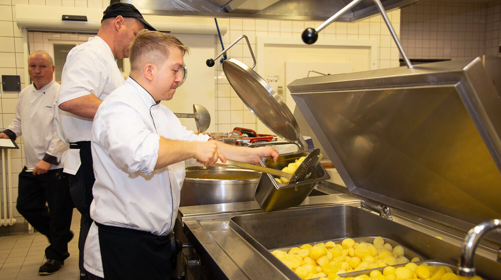 Ein Mann in weißer Arbeitskleidung füllt in der Großküche gekochte Kartoffeln in eine kleinere Box