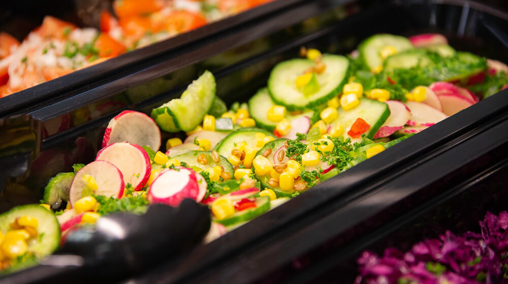 Eine längliche Salatschale auf einem Buffet ist mit buntem Salat gefüllt und eine Zange liegt in ihr, um sich zu bedienen.
