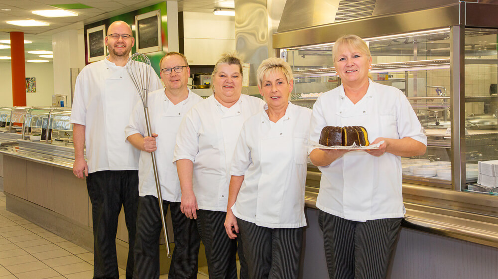 Fünf Mitarbeitende in Arbeitskleidung stehen zusammen und halten Kochgeschirr und einen gebackenen Kuchen