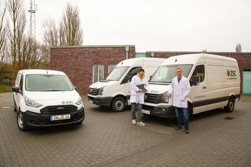 Drei Transportautos stehen nebeneinander und zwei Mitarbeiter lehnen sich an eines an.