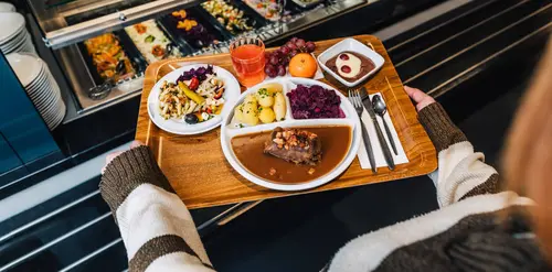 Eine Person hält ein Tablett mit Hauptspeise, Salat und Dessert