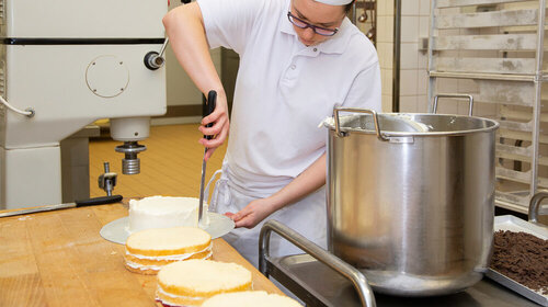 Eine Frau mit Haube und in Bäckerkleidung verziert eine Torte mit einem Konditormesser