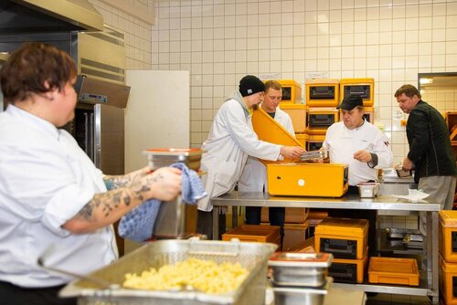 In einer Großküche portionieren Mitarbeitende Speisen in große Gefäße und Transportboxen.
